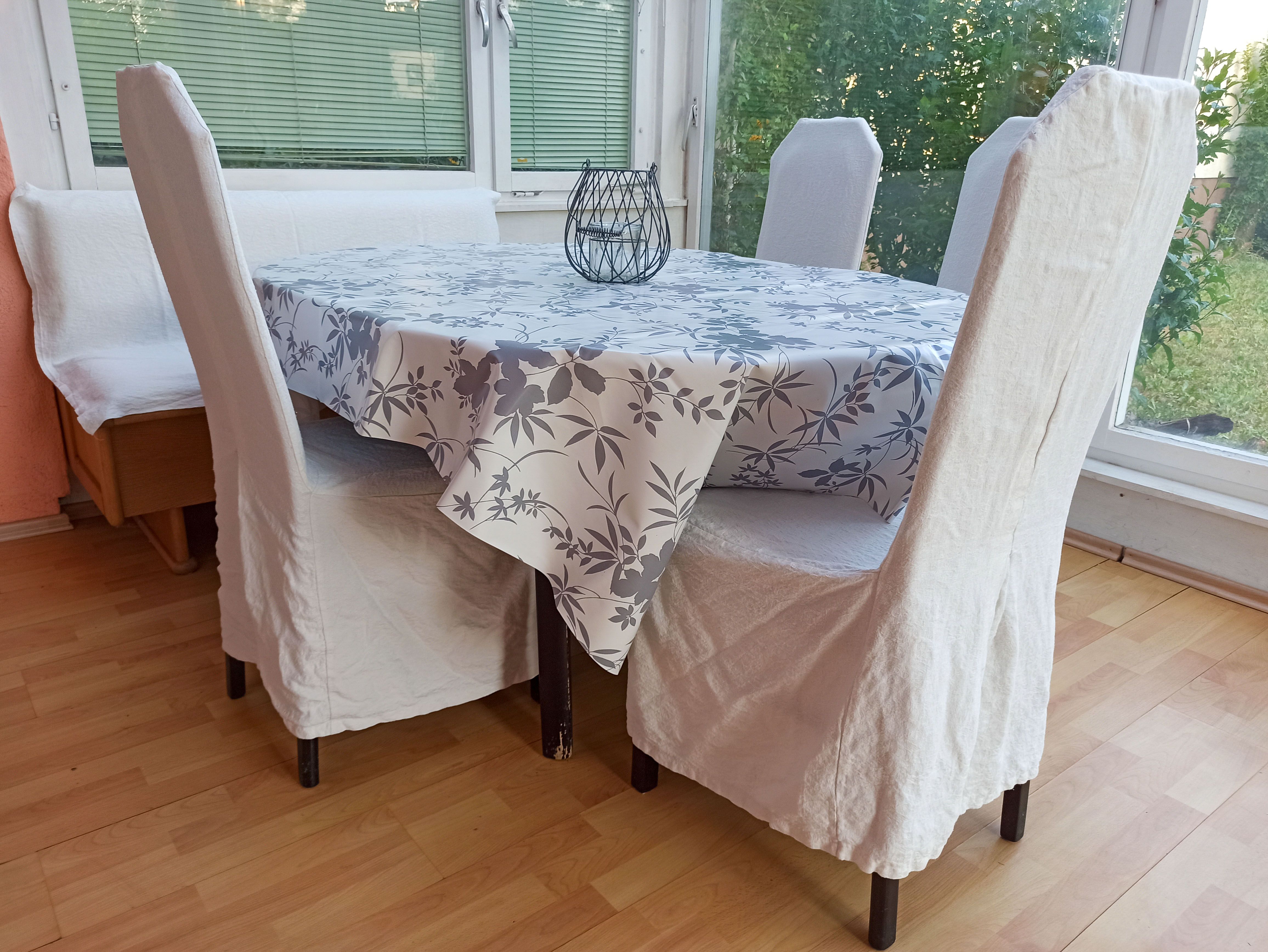 Dining area with table, chairs and bench, tablecloth with floral pattern, window in the background.