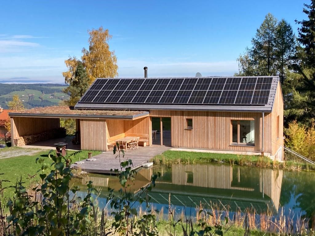 A modern wooden house with solar panels on the roof, surrounded by nature and a small pond in the foreground.