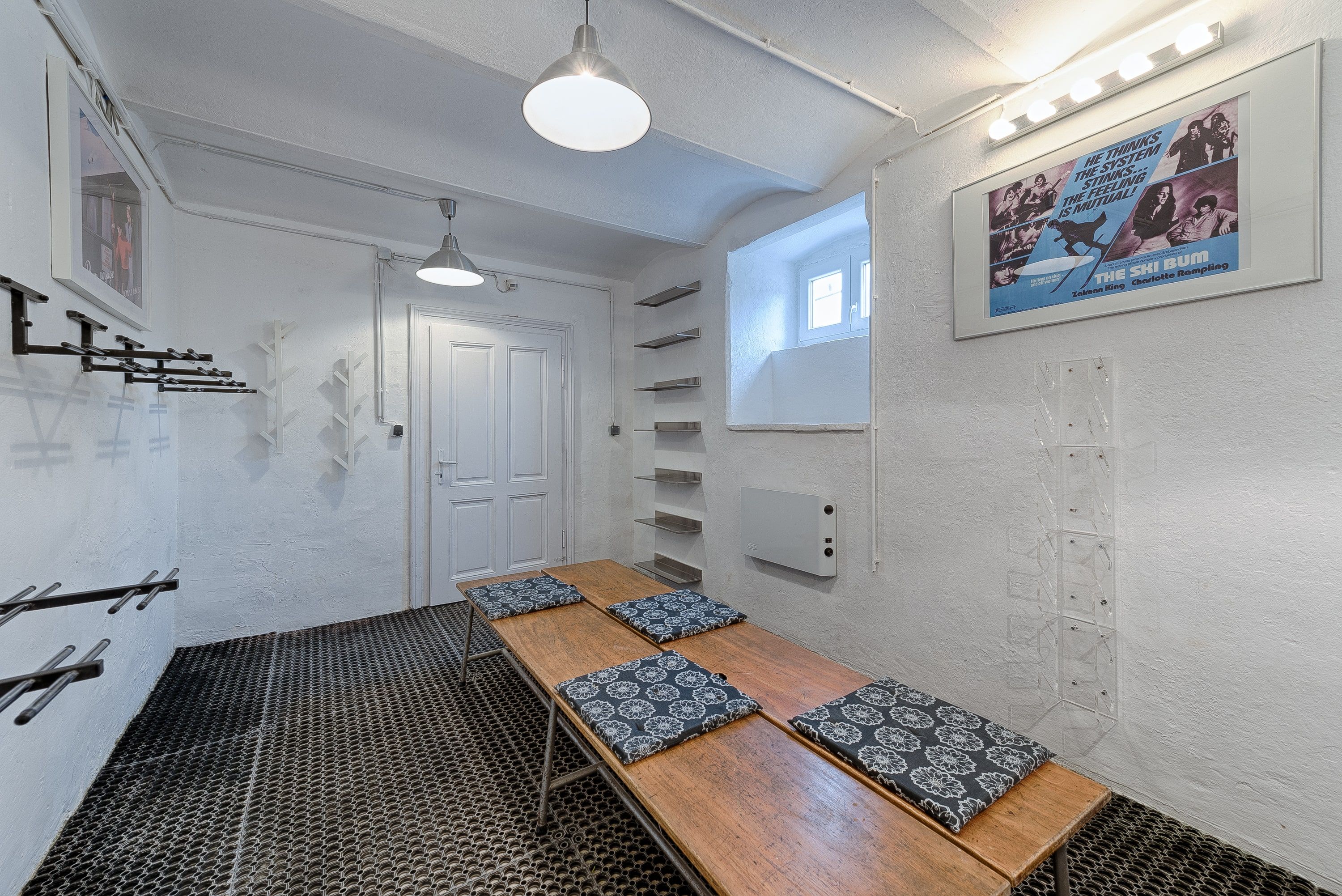 A ski cellar with white walls, ski racks, a wooden bench with cushions and a movie poster on the wall.