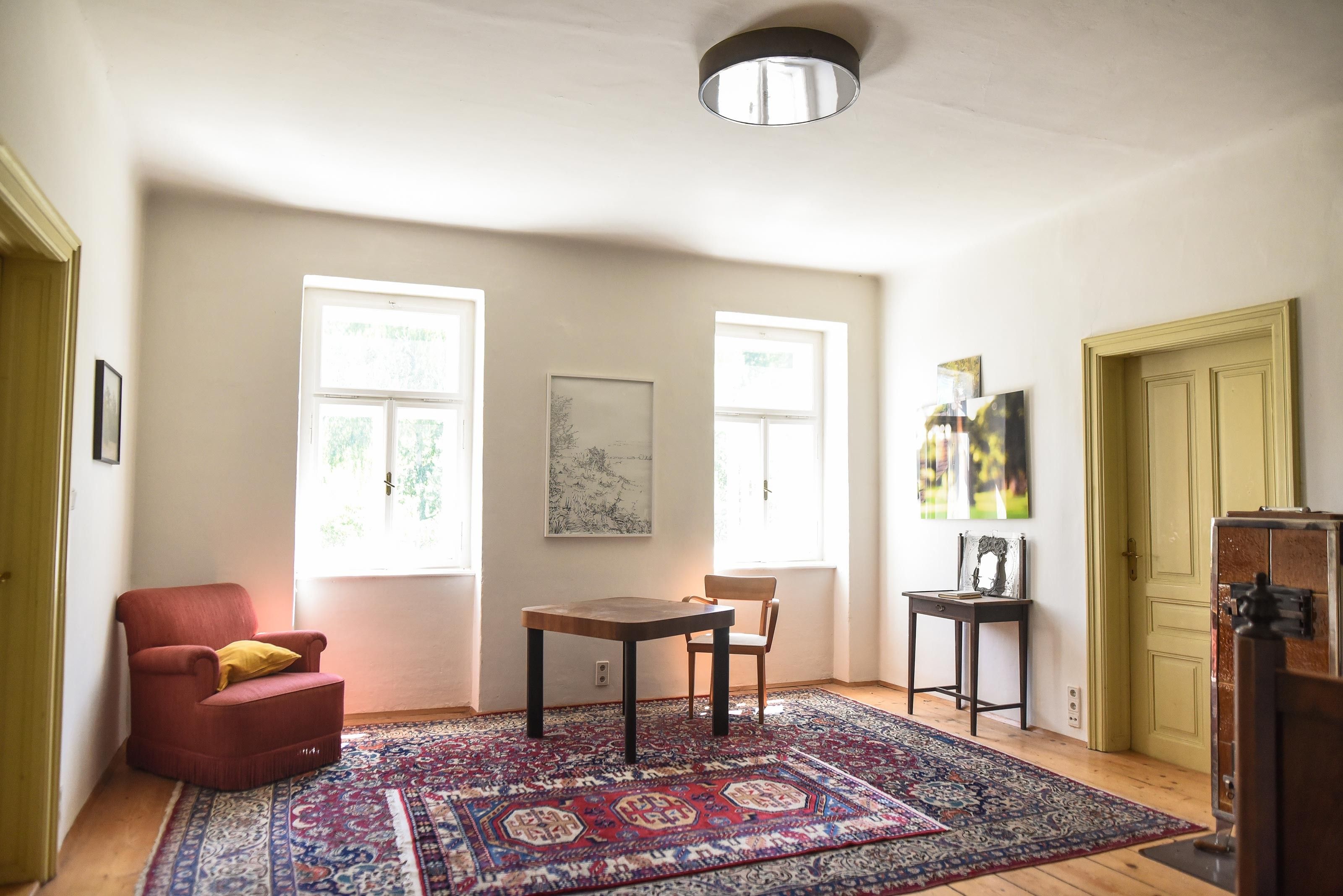 A bright living room with a red armchair, a wooden table, and a chair on an oriental rug.