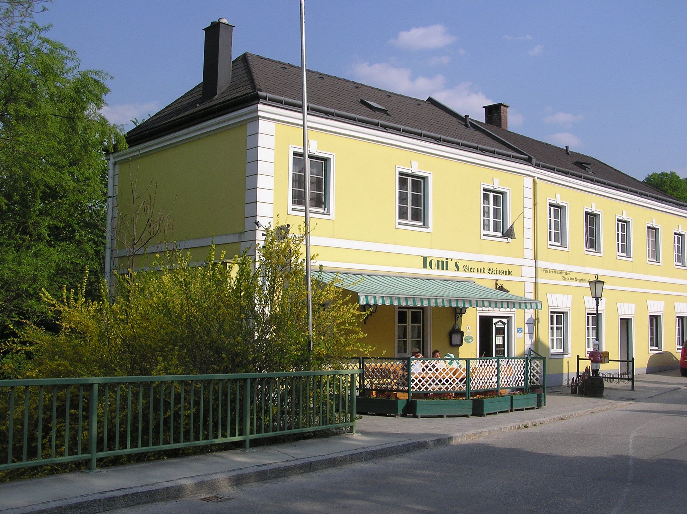Yellow building with the inscription 'Toni's Bier- und Weinstube', surrounded by green plants and a fence.