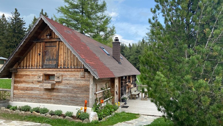A rustic wooden hut with a red roof in a wooded setting, surrounded by trees and a well-tended garden.
