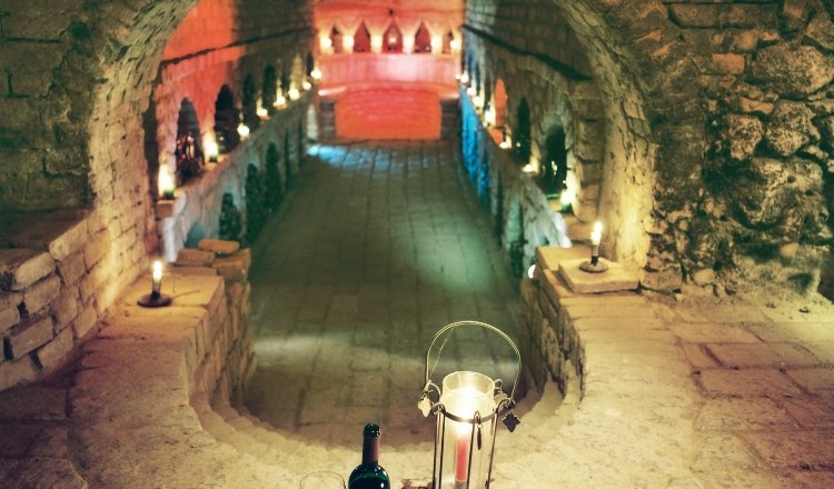 An atmospherically lit wine cellar with candles, a bottle of wine and a glass on a stone ledge.