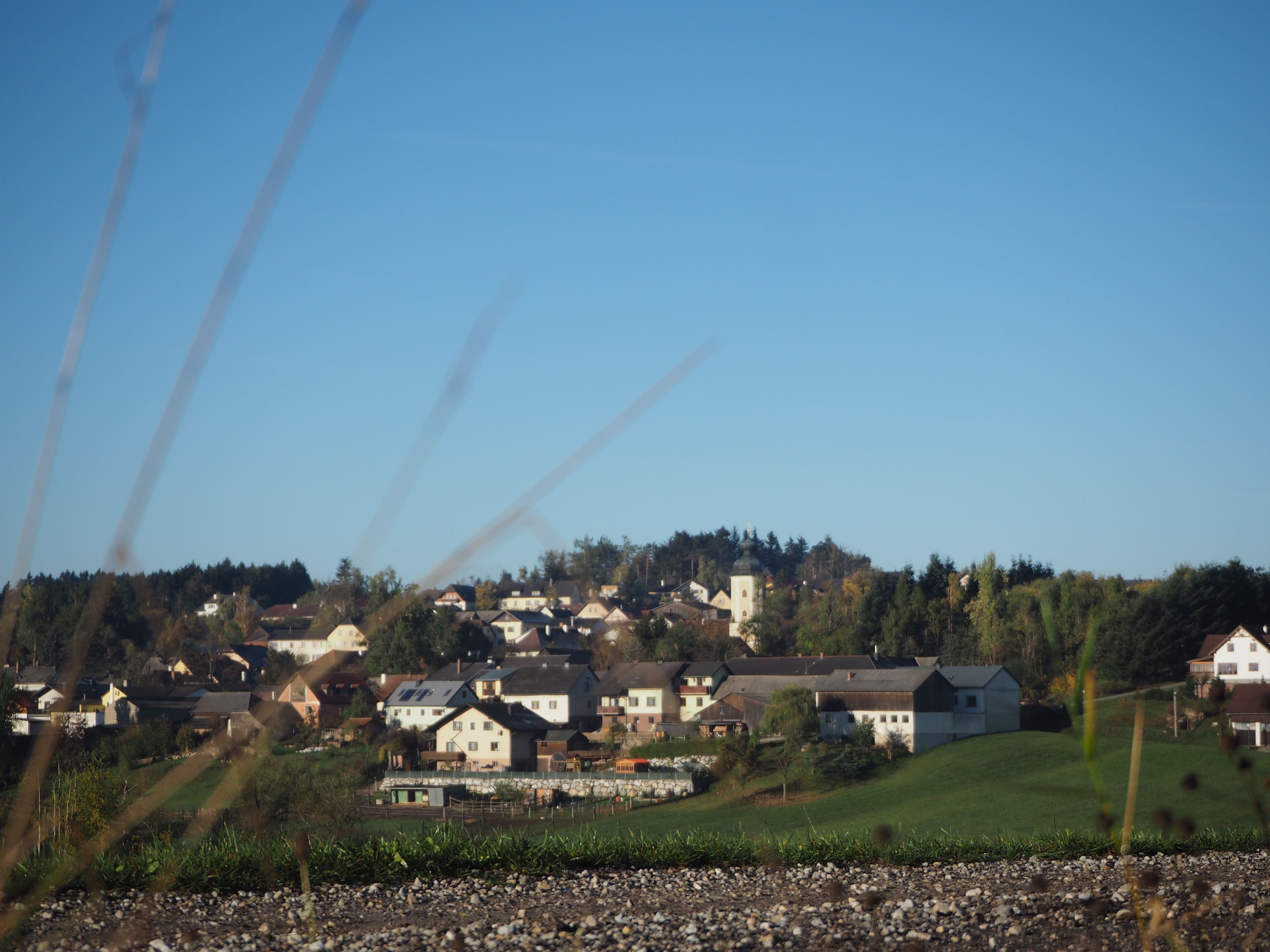 Rural settlement with church and houses on a hill, surrounded by trees and meadows under a clear sky.