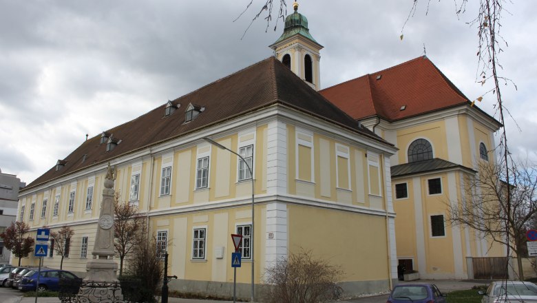 Historic building with a yellow façade and red roof, next to a statue and parked cars.