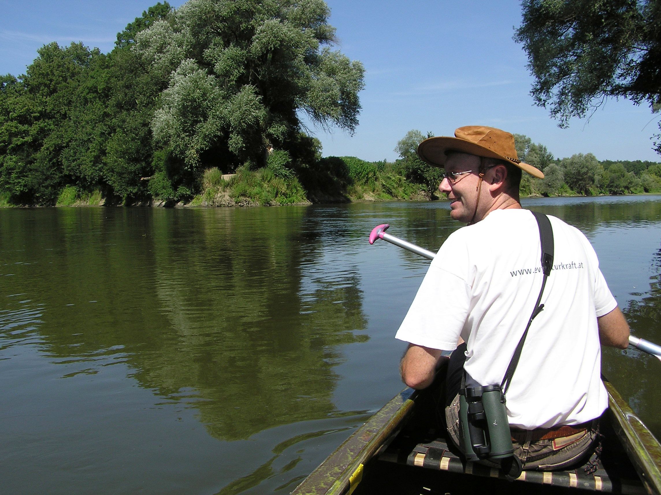 A man in a hat paddles in a canoe on a river surrounded by green trees.