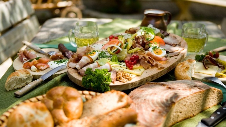 A richly laid table with a selection of cold meats, cheese, bread and beer mugs outside.
