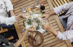 Three people clink glasses of wine at a wooden table with bread, water and flowers.