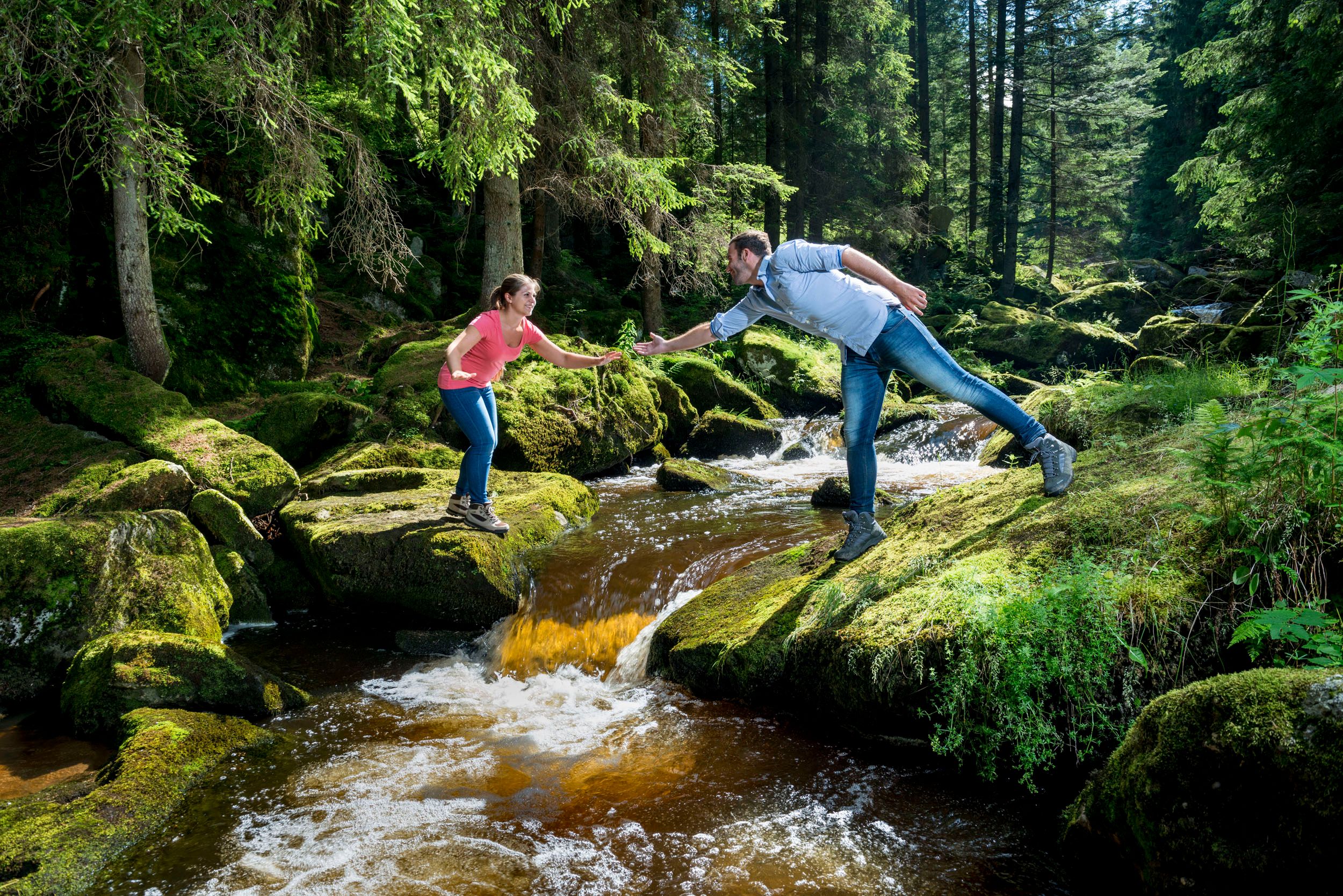 Two people stand on moss-covered stones above a stream in a forest and shake hands.