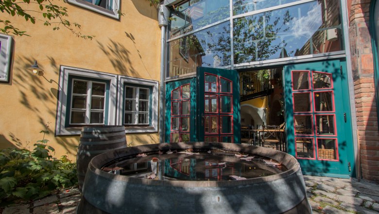 An inner courtyard with a yellow façade, green doors and a wooden barrel of water. Shadows of trees fall on the wall.