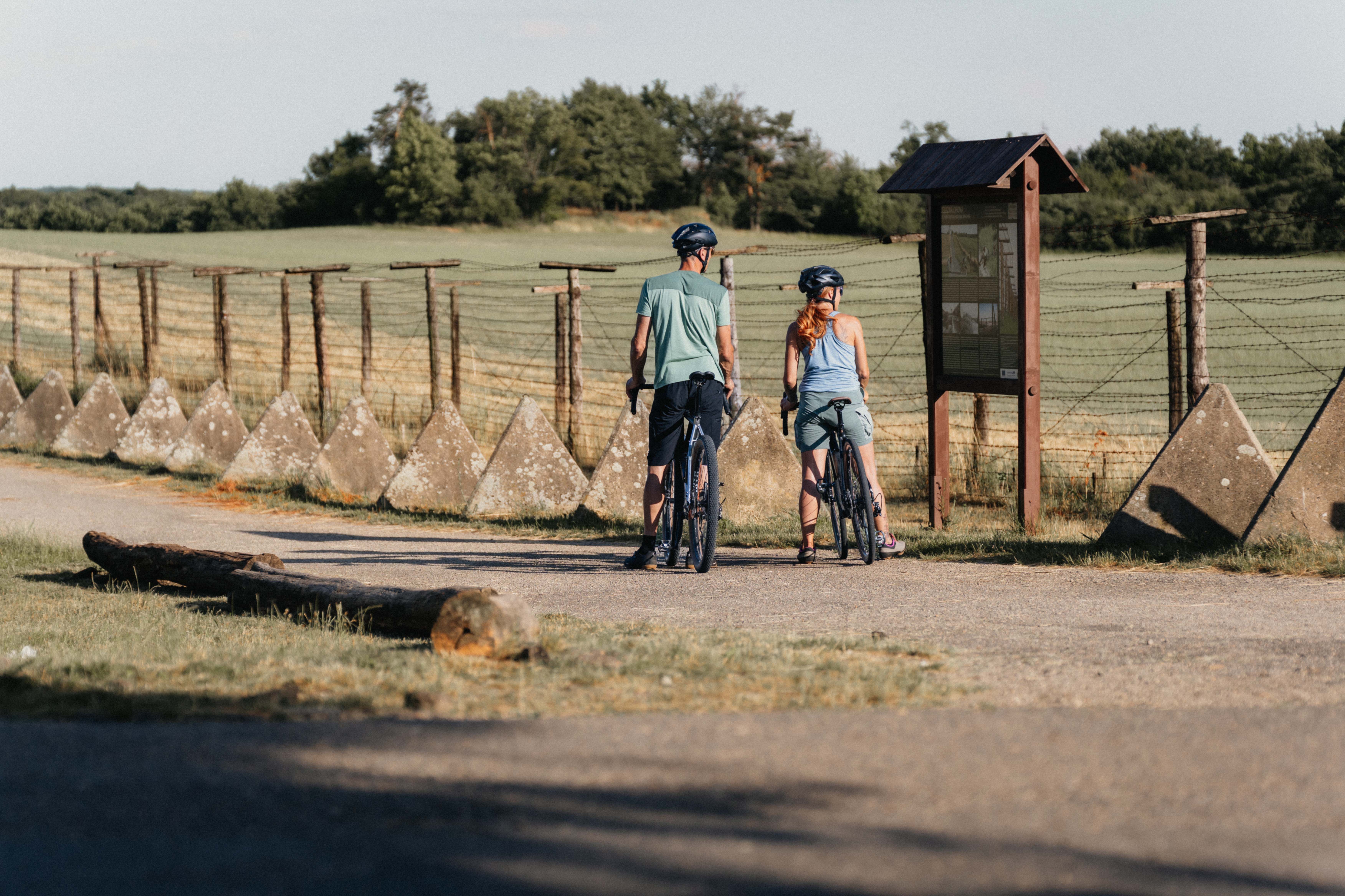 Two cyclists stand in front of a historic border fence with an information board.