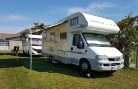 A white motorhome with an extended awning is parked on a meadow next to another vehicle.