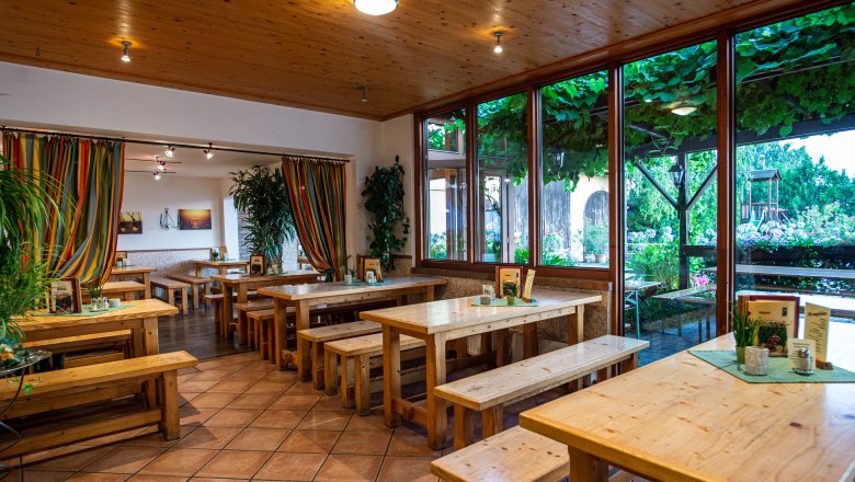 Interior view of a wine tavern with wooden tables and benches, colorful curtains and large windows overlooking a leafy outdoor area.