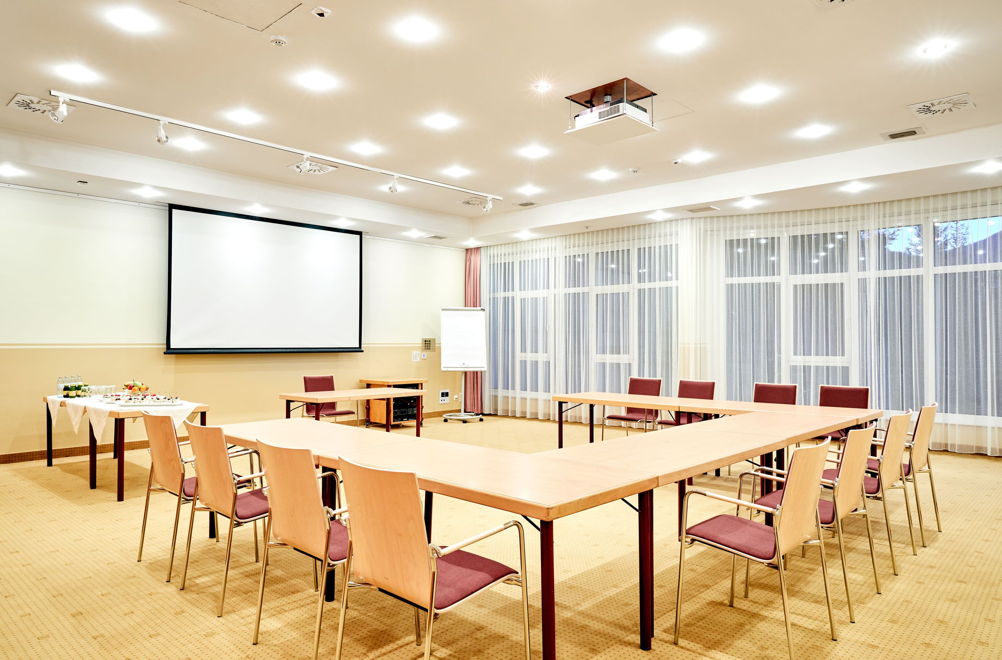 A bright seminar room with a U-shaped table arrangement, screen, flipchart and drinks table.
