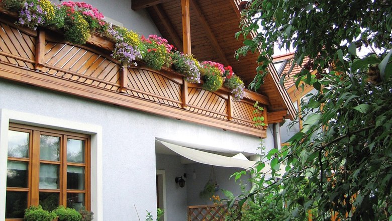 A guest house with a wooden balcony and blooming flowers, surrounded by green plants.