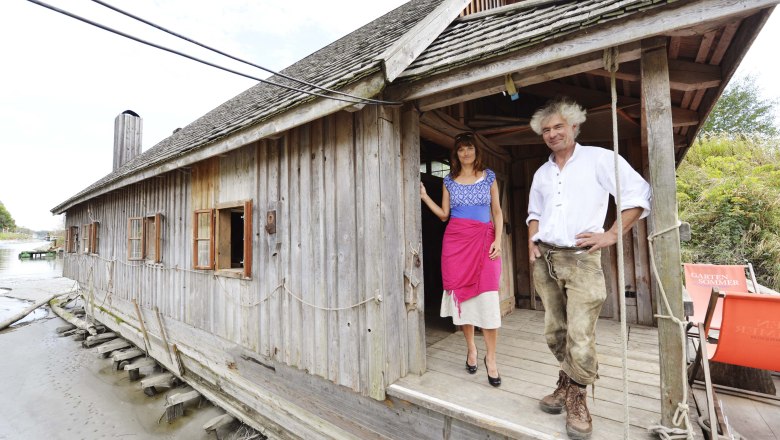 Two people are standing on the veranda of a wooden ship's mill on a riverbank.