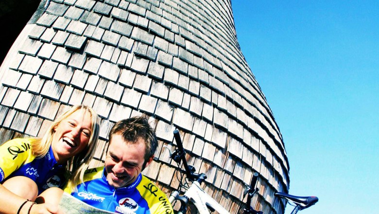 Two cyclists sit in front of a large pear-shaped wooden structure and laugh.