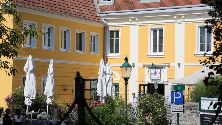 Exterior view of a yellow building with red roof tiles and white window frames, surrounded by plants and parasols.