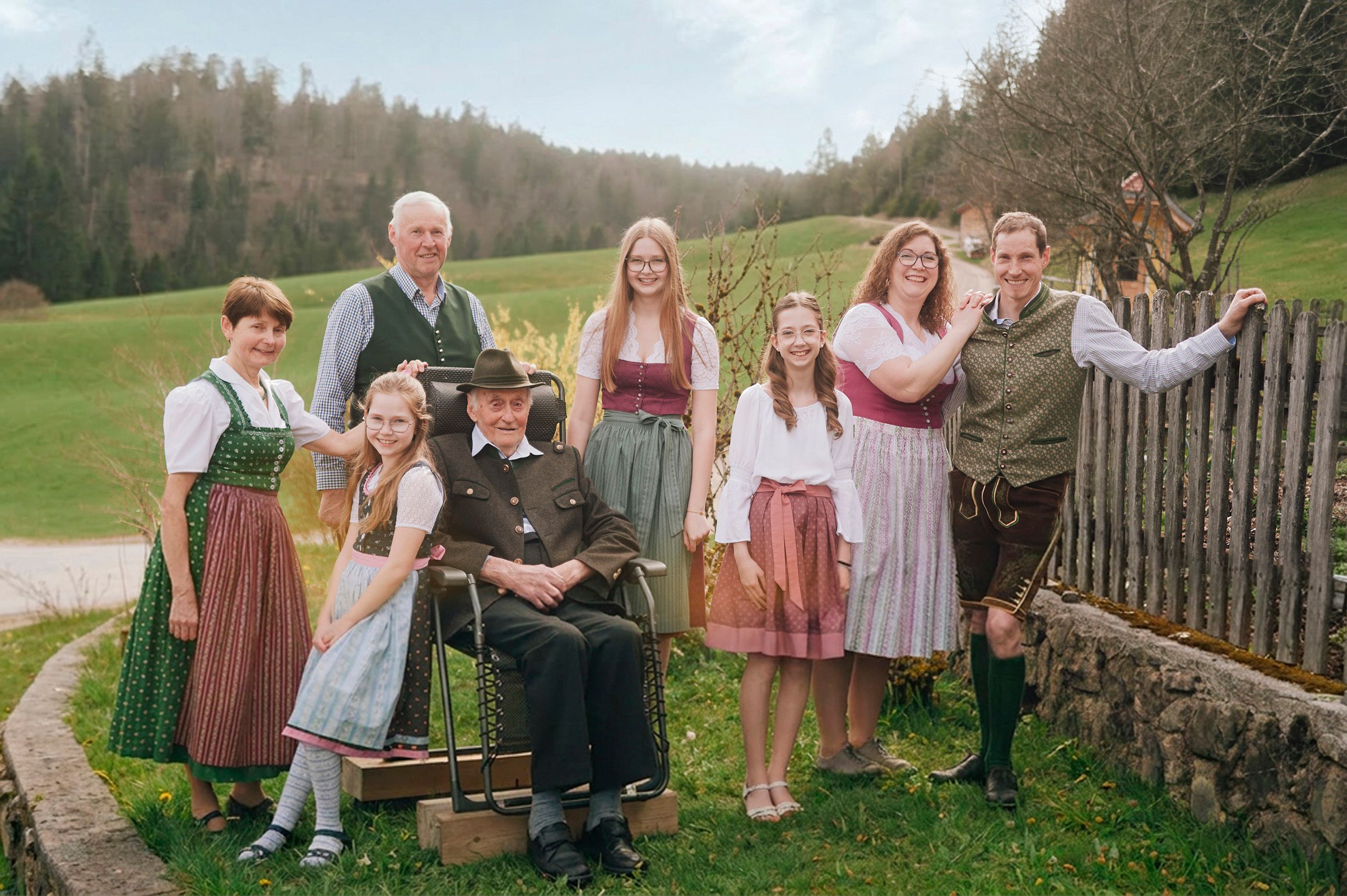 A family in traditional costume poses outdoors against a rural backdrop of meadows and trees.