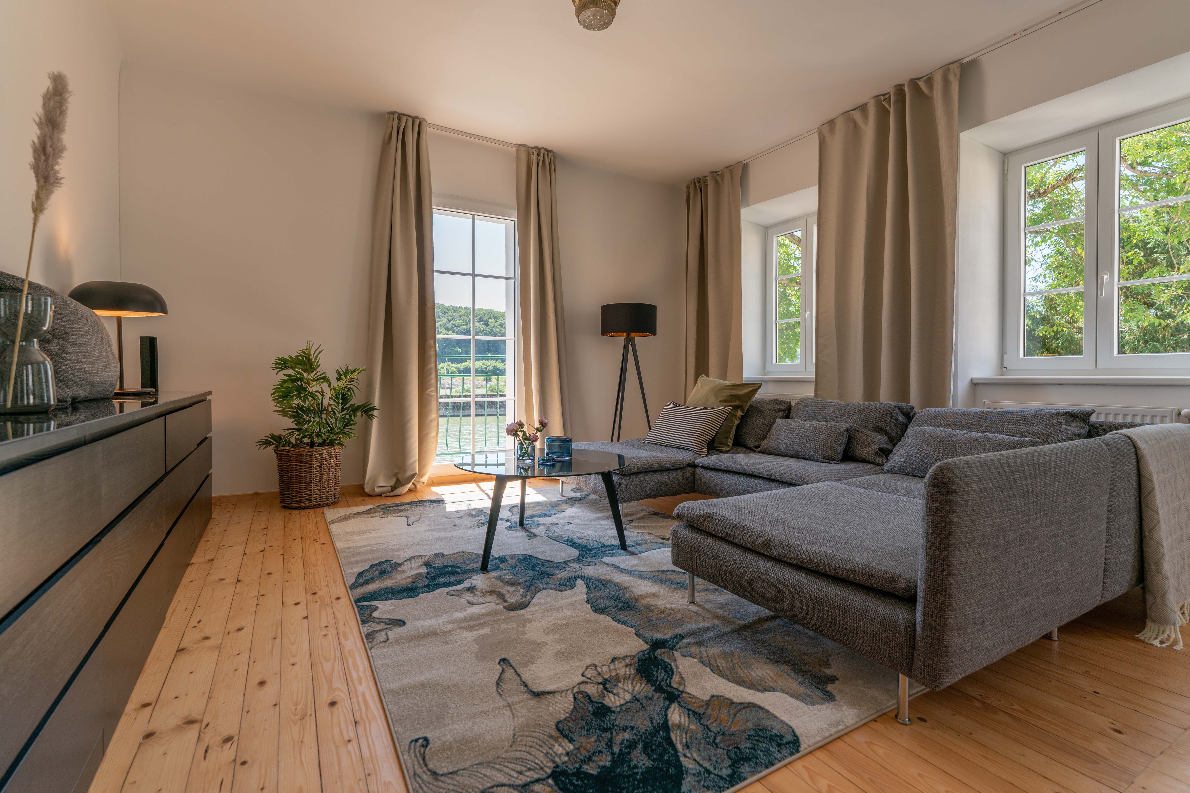 Modern living room with gray sofa, wooden floor and large window with a view of the greenery.