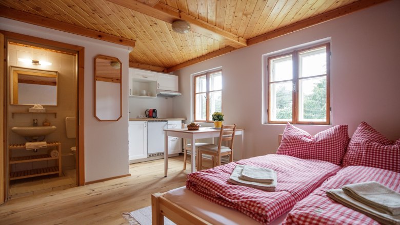Cozy vacation apartment with wooden floor, double bed, small kitchen and dining table. Bathroom in the background.