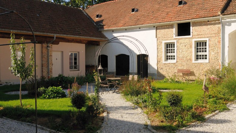 Inner courtyard, © Franz Böck An idyllic inner courtyard with gravel paths, green plants and a seating area under a metal arch.