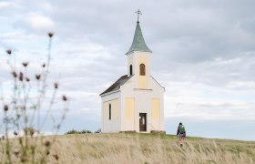 A small chapel on a hill with a person walking towards it, surrounded by a meadow and a cloudy sky.