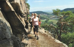 Three people hike on a narrow path along a rock face with a view of a green landscape.