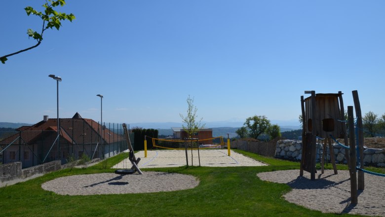 A beach volleyball court with a net, surrounded by green grass and a playground in the foreground, under a clear blue sky.