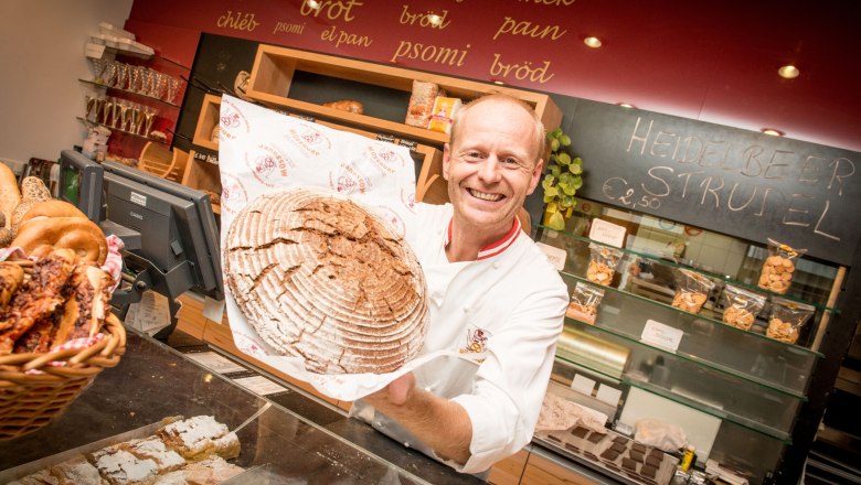 A baker in a bakery holds a large, round loaf of bread up to the camera with a smile. In the background are shelves of baked goods and a board with the inscription 'Heidelbeer Strudel'.