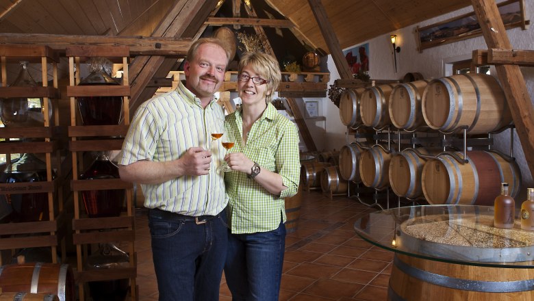 A couple is standing in a distillery with wooden barrels and holding glasses of spirits.