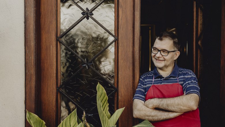 A man with glasses and a red apron stands smiling in a doorway next to a window with a wooden frame and a plant.