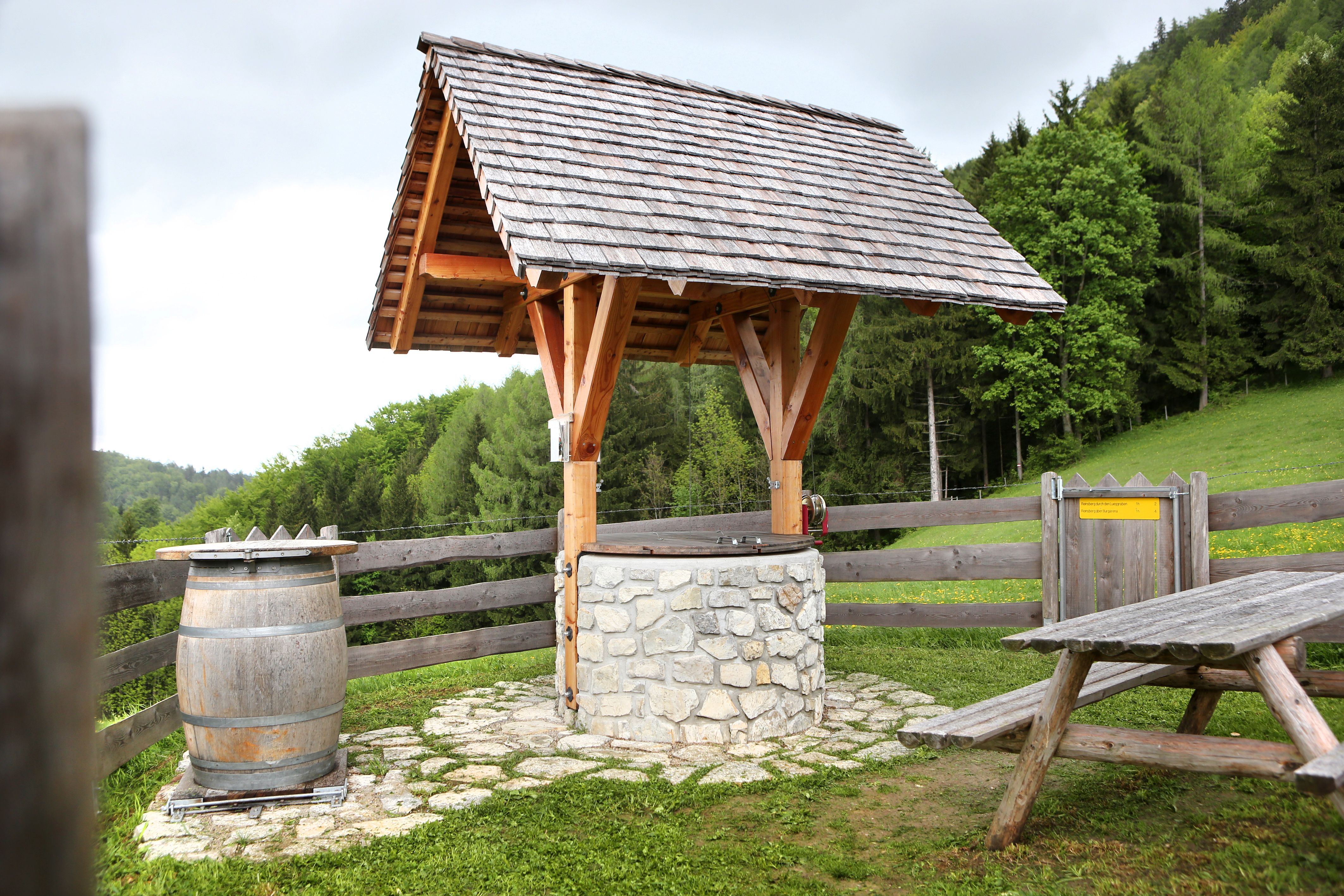 A traditional well with a wooden roof and stone wall, next to a wooden barrel and a picnic bench, surrounded by green countryside.