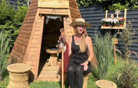 Woman in a straw hat sits in front of a wooden tepee in the garden.