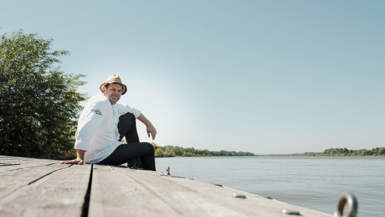 Man in a hat and chef's jacket sitting on a wooden jetty on the riverbank, sunny day.