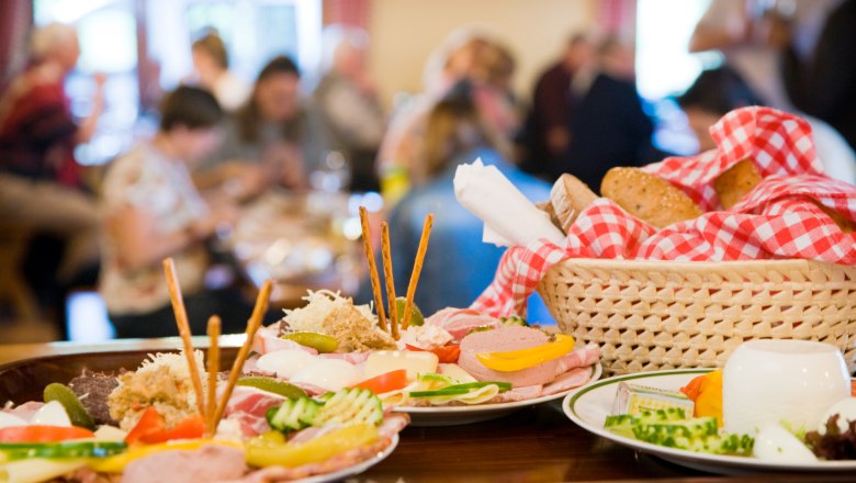 A table with traditional Austrian dishes, including cold cuts, cheese and bread in a basket with a red and white checked cloth.