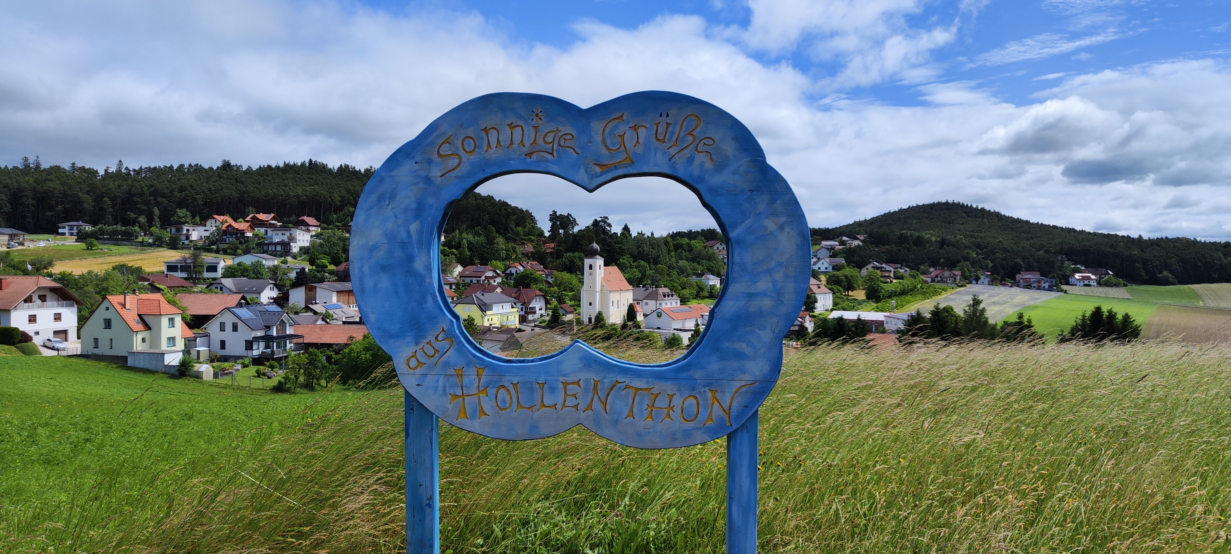 View through a blue frame with the inscription 'Sunny greetings from Hollenthon' to a picturesque landscape with village and church.