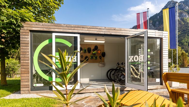 A modern bike box with open doors, bikes and helmets, surrounded by nature and flags.