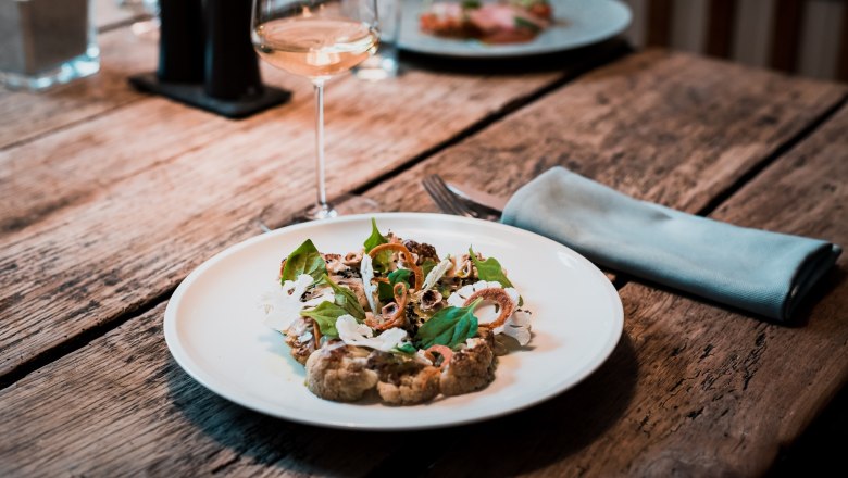 A plate of braised cauliflower, miso spinach and pickled sea buckthorn on a wooden table, with a glass of white wine next to it.
