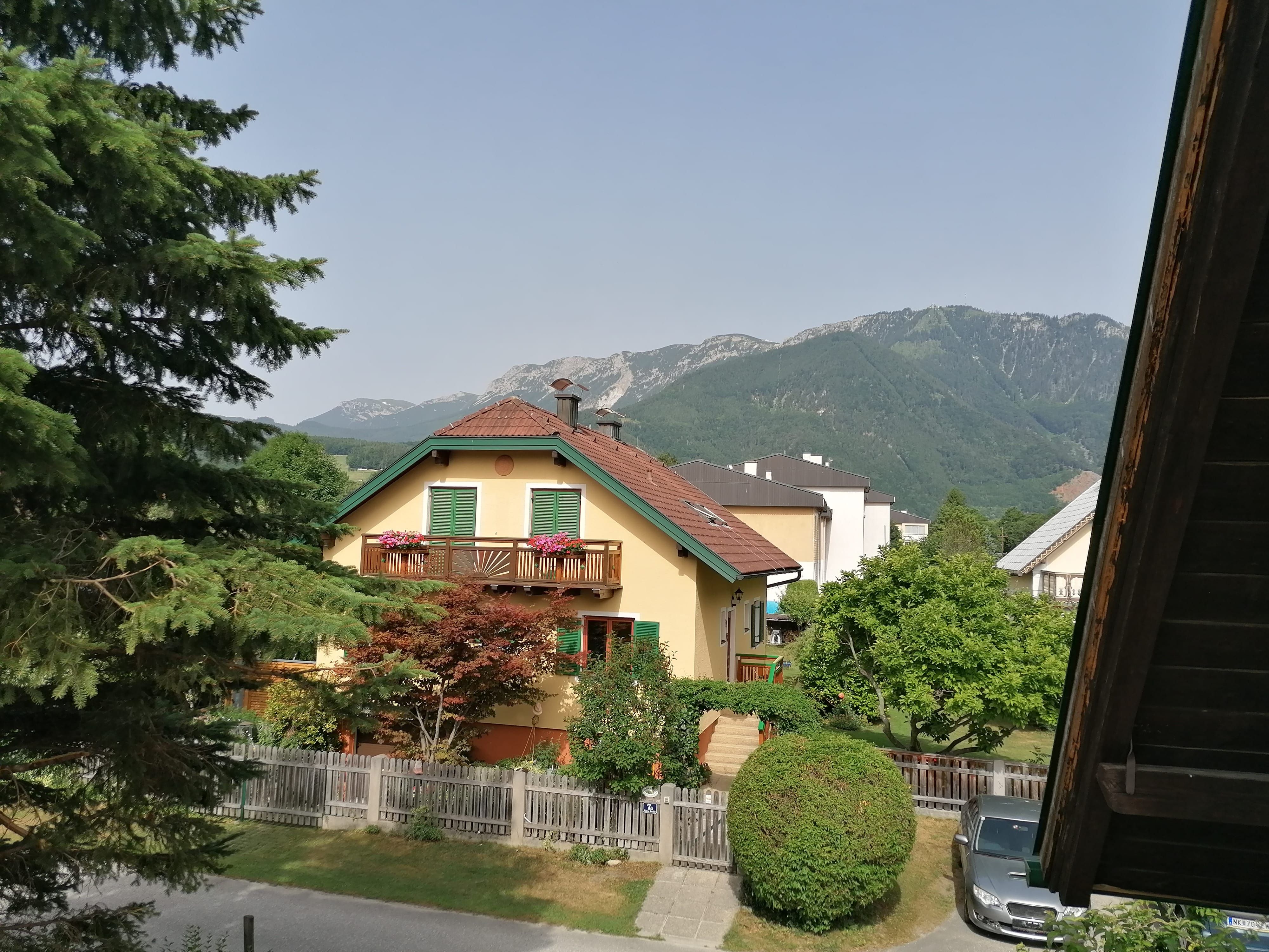 View from a balcony of a yellow house with a red roof, surrounded by trees and mountains in the background.