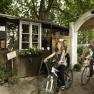 Three people on bicycles ride through an archway in a garden with chestnut trees.
