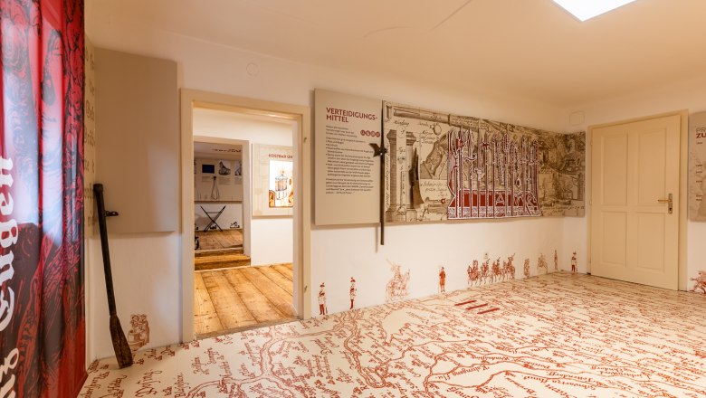 Interior view of a museum room with historical maps and weapons on the walls, a large old map is drawn on the floor