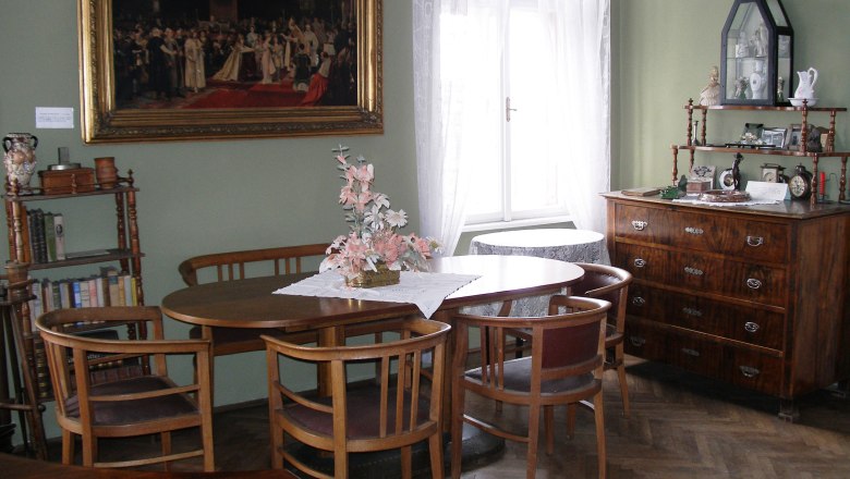 A traditionally furnished living room with wooden furniture, a round table, chairs, a chest of drawers and a large painting on the wall.