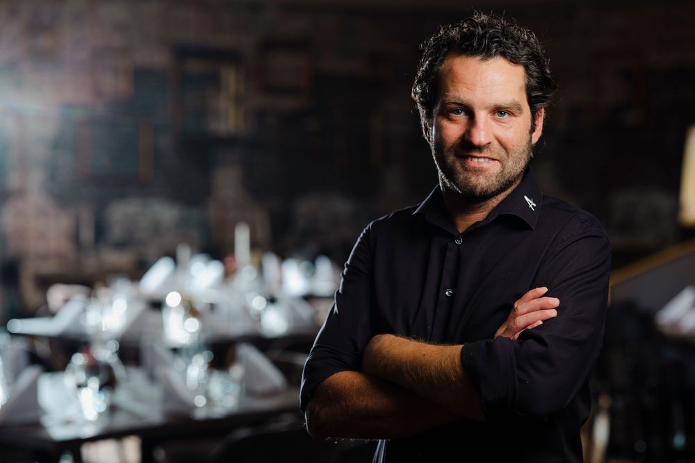 Man in a black shirt stands in front of an elegantly laid table in a dark room.