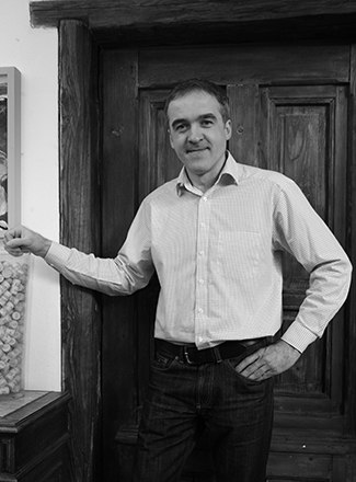 A man in a shirt stands in front of a wooden door and smiles at the camera.