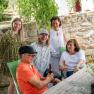 Group of five people sitting and standing at an outdoor table, surrounded by plants and a stone wall.
