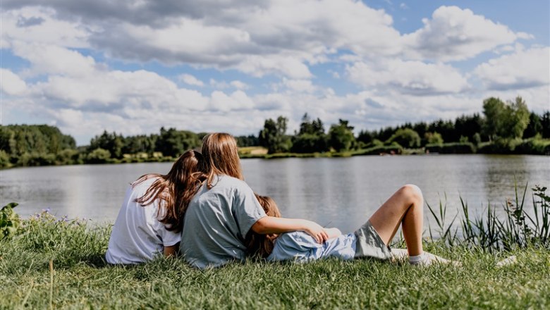 Three people sit on a meadow by the lake and look out over the water.