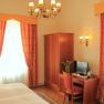 A cozy hotel room with a double bed, wooden furniture, a desk with TV and a vase of flowers. Curtains in red-orange.