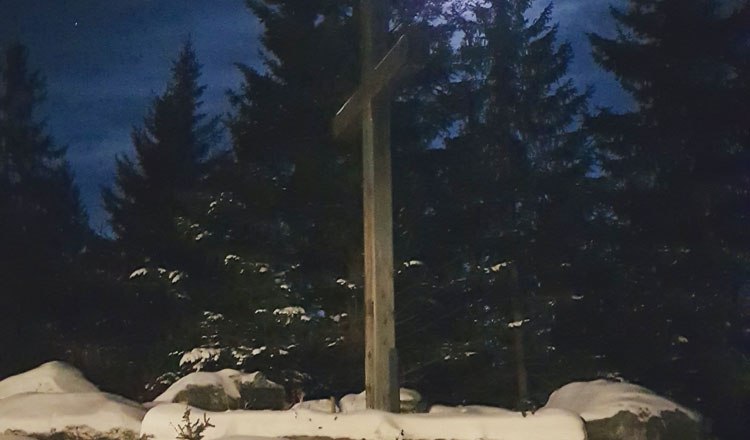 Snow-covered benches and tables in front of a cross, illuminated by the full moon, surrounded by fir trees.