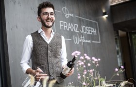Man with glasses and vest holding wine bottle in front of restaurant 'Franz Joseph'.